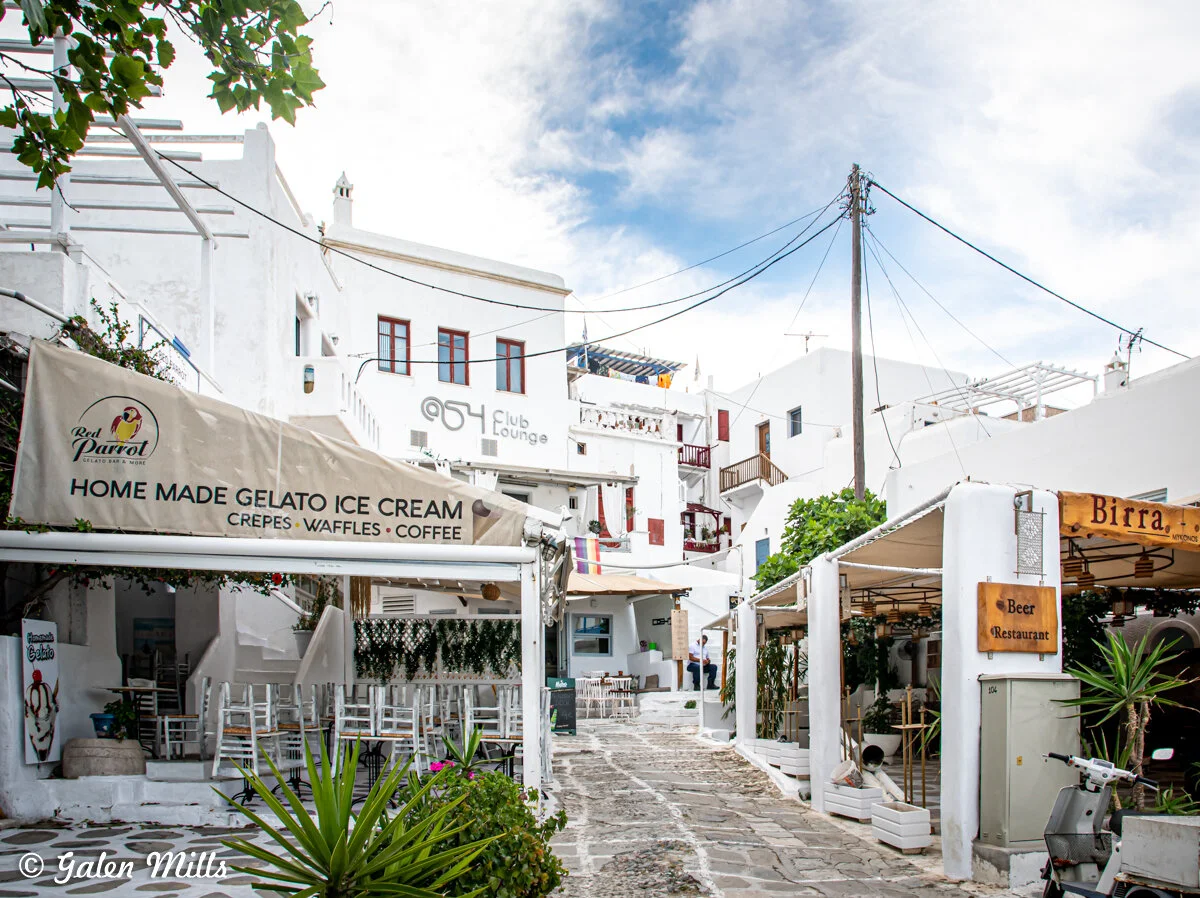 White-washed buildings in a quaint narrow street with cafes and restaurants in Mykonos, Greece, featuring signs for homemade gelato and a club lounge.