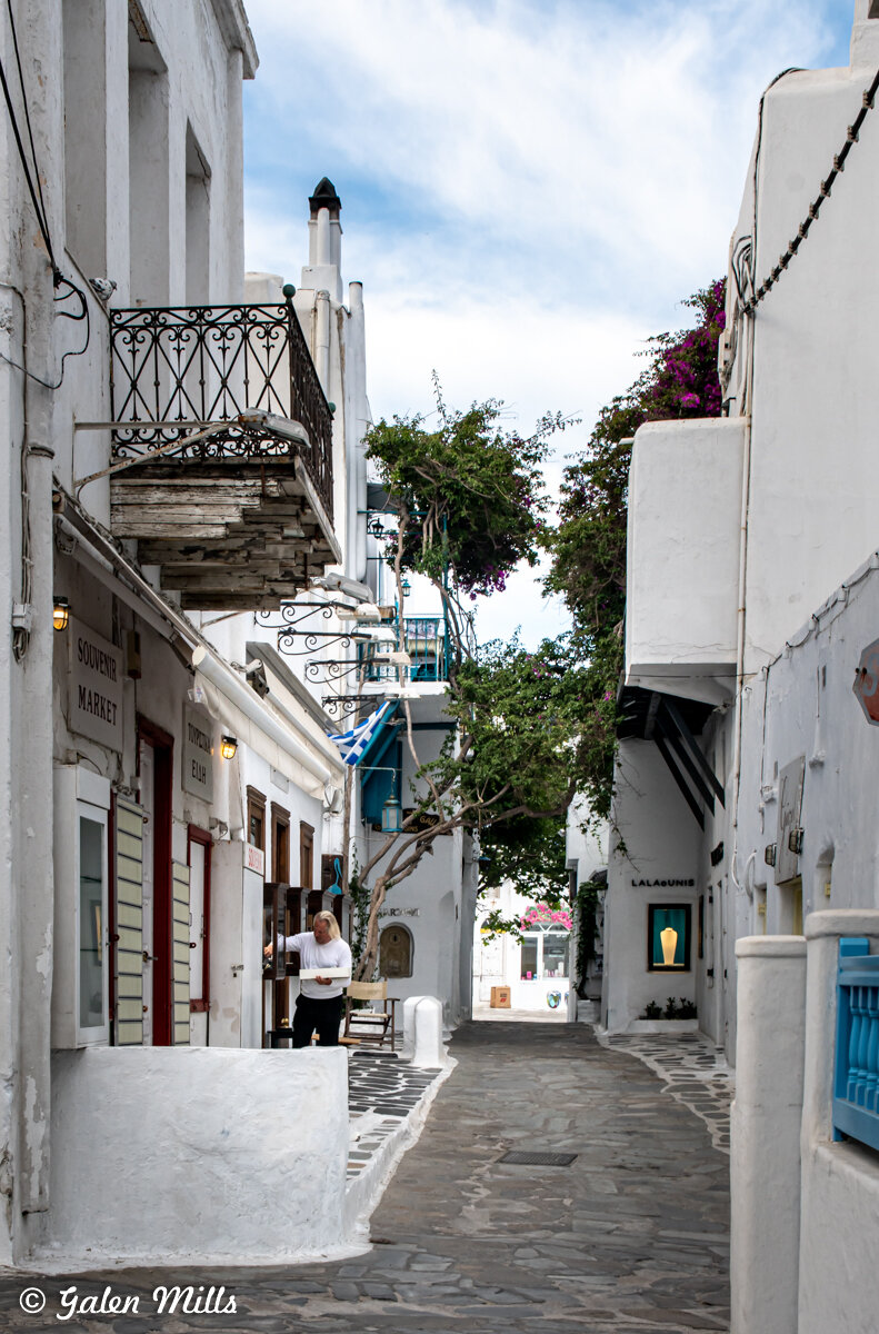Narrow cobblestone street in Mykonos, Greece, lined with white buildings and shops. A person is standing near a doorway. A small balcony and flowering trees enhance the scene.