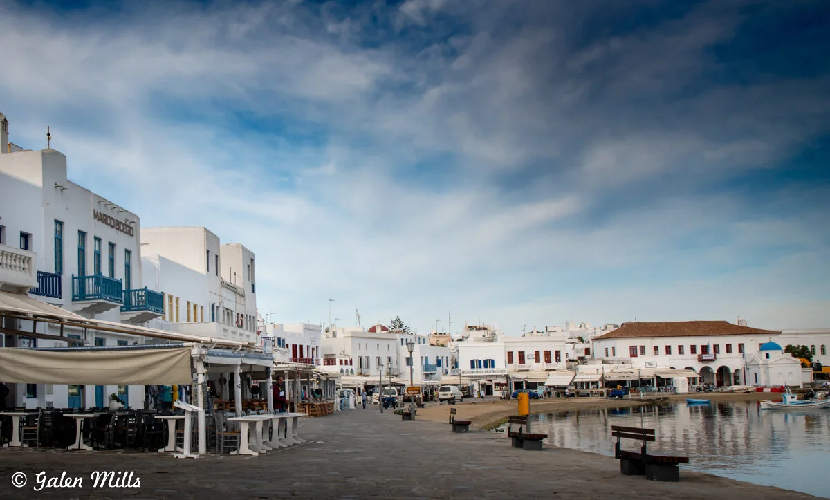 Waterfront with white buildings and blue accents in Mykonos, Greece, under a cloudy sky.