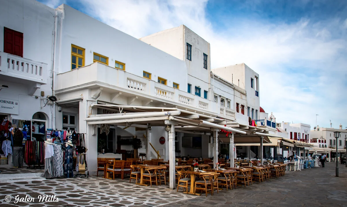 Outdoor café and shops on a cobblestone street with white buildings and colorful windows in a Mediterranean town.