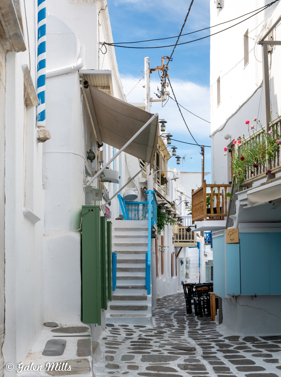 Narrow cobblestone street in a Greek island village with whitewashed buildings, green and blue shutters, outdoor seating, and flowers.