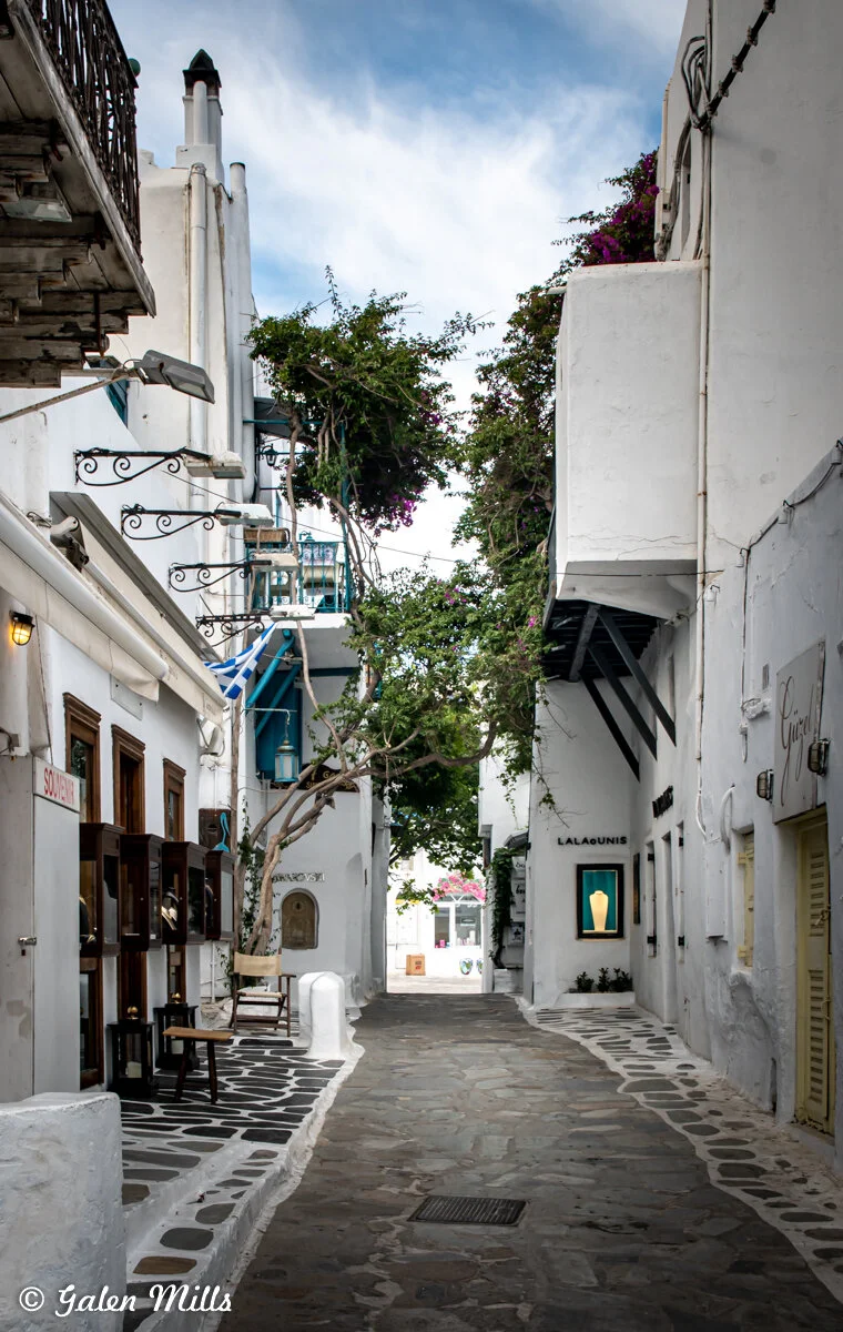 Narrow street in Mykonos, Greece with whitewashed buildings, cobblestone path, and blooming bougainvillea.