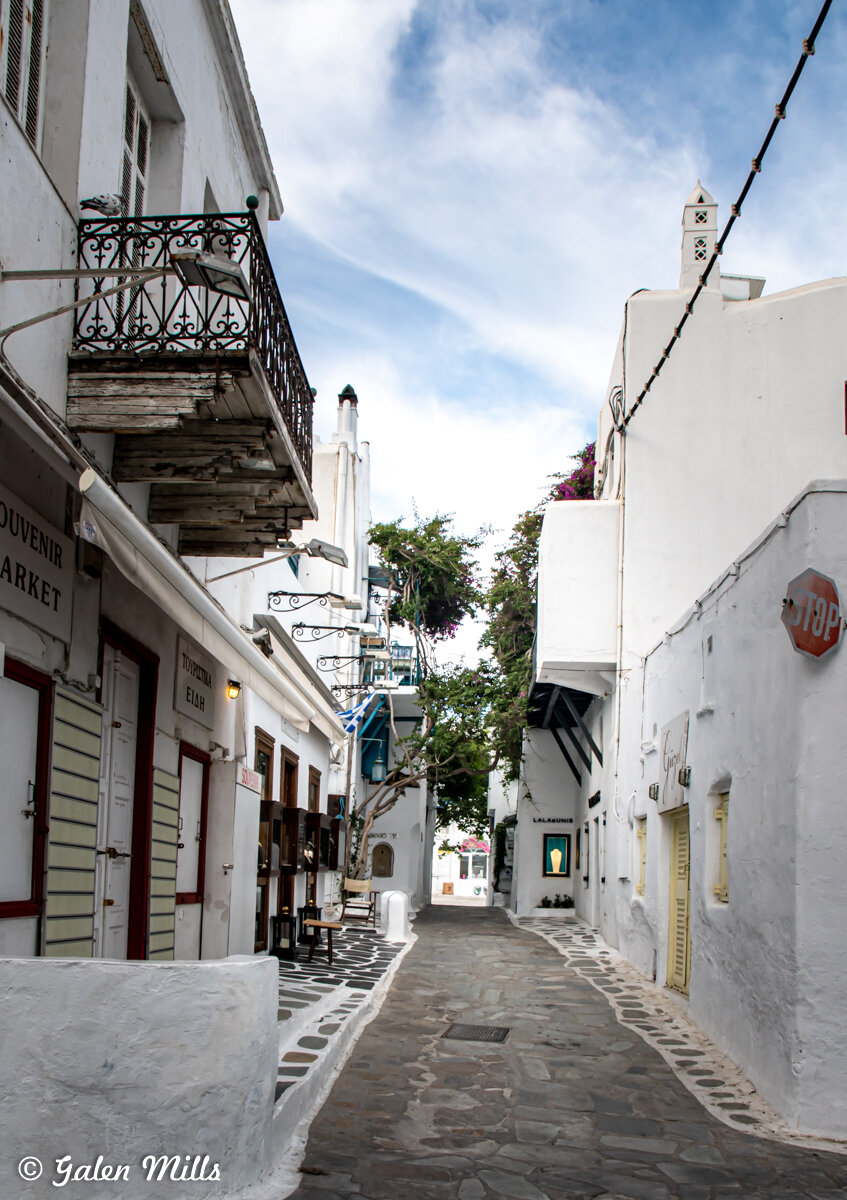 Narrow cobblestone street in Mykonos, Greece, lined with traditional whitewashed buildings and ornate balconies, under a clear blue sky.