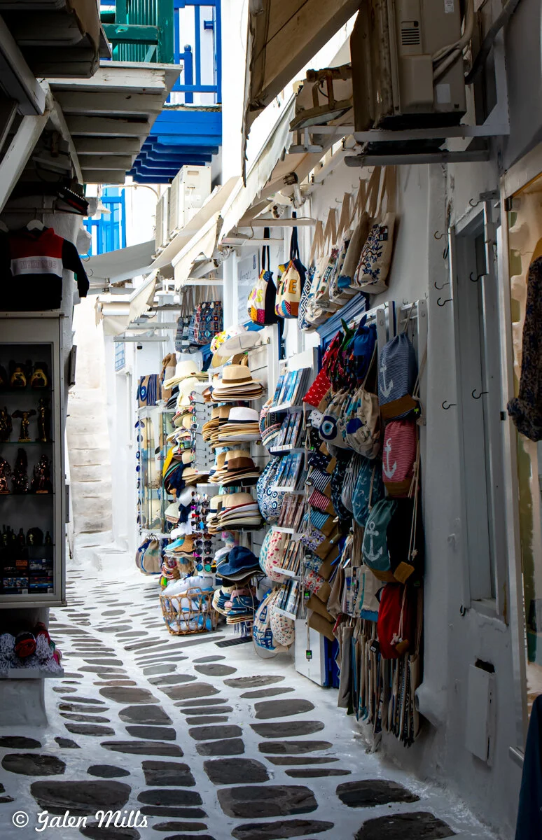 Narrow street lined with shops displaying hats, bags, and souvenirs.