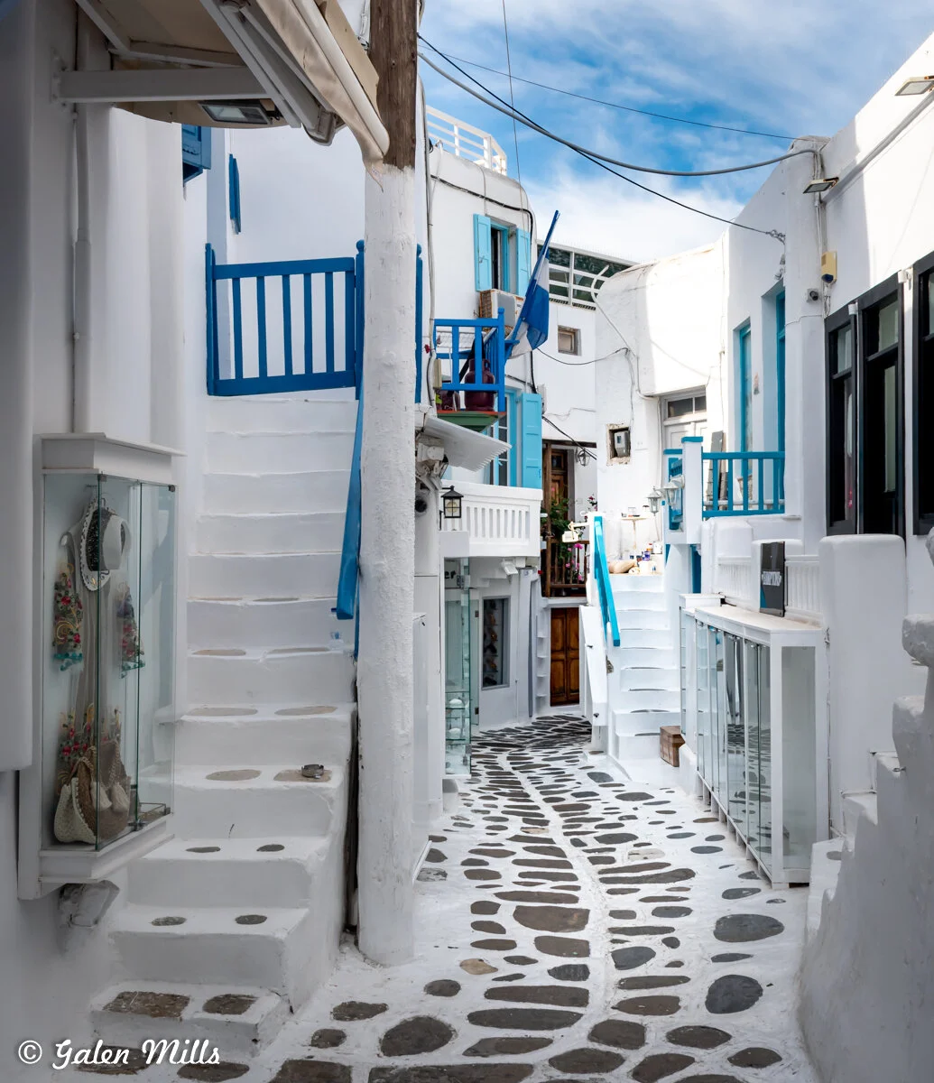 Narrow alley in Mykonos, Greece, with whitewashed buildings, blue doors and windows, and stone-paved pathway.