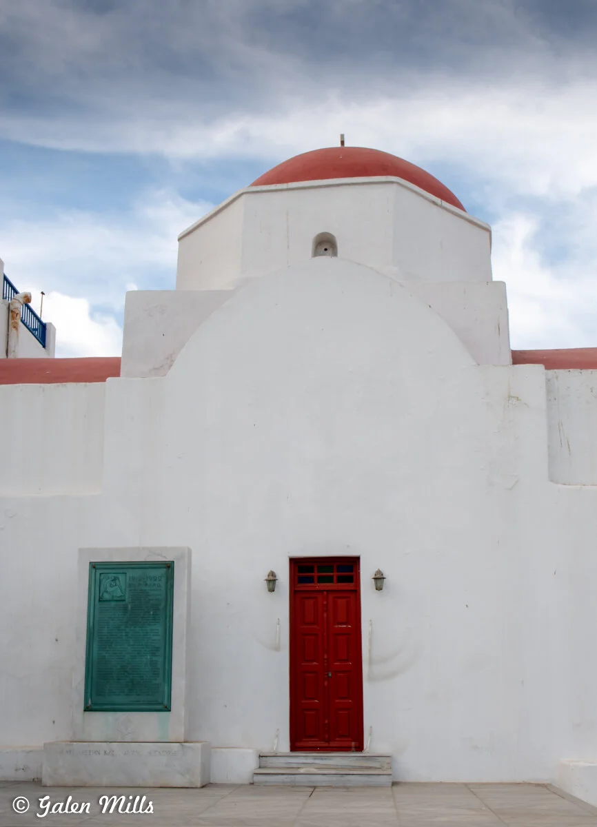 White building with a red dome and door, featuring a plaque, in front of a cloudy sky.
