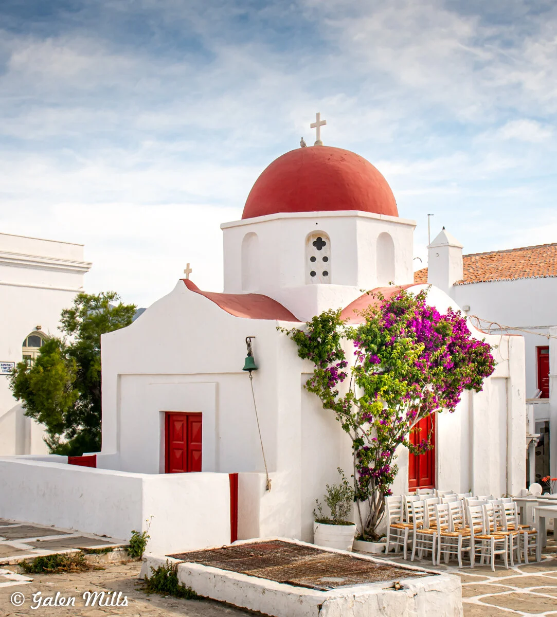 Traditional Greek white chapel with a red dome, adorned with a cross on top, surrounded by vibrant purple bougainvillea and white patio furniture, set against a blue sky.