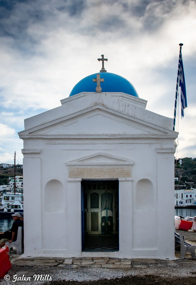Small white chapel with a blue dome and cross, Greek flag, cloudy sky.