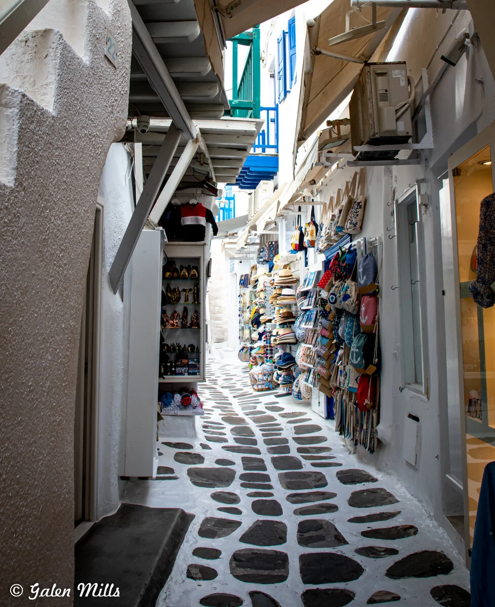 Narrow alley in Mykonos, Greece, with whitewashed walls and stone path, lined with shops displaying hats, bags, and souvenirs.