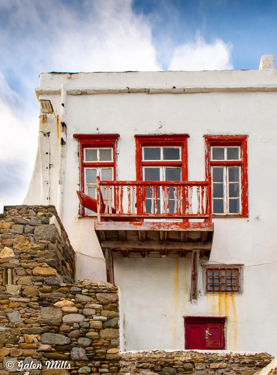 White building with red-trimmed windows and balcony, stone wall, blue sky.