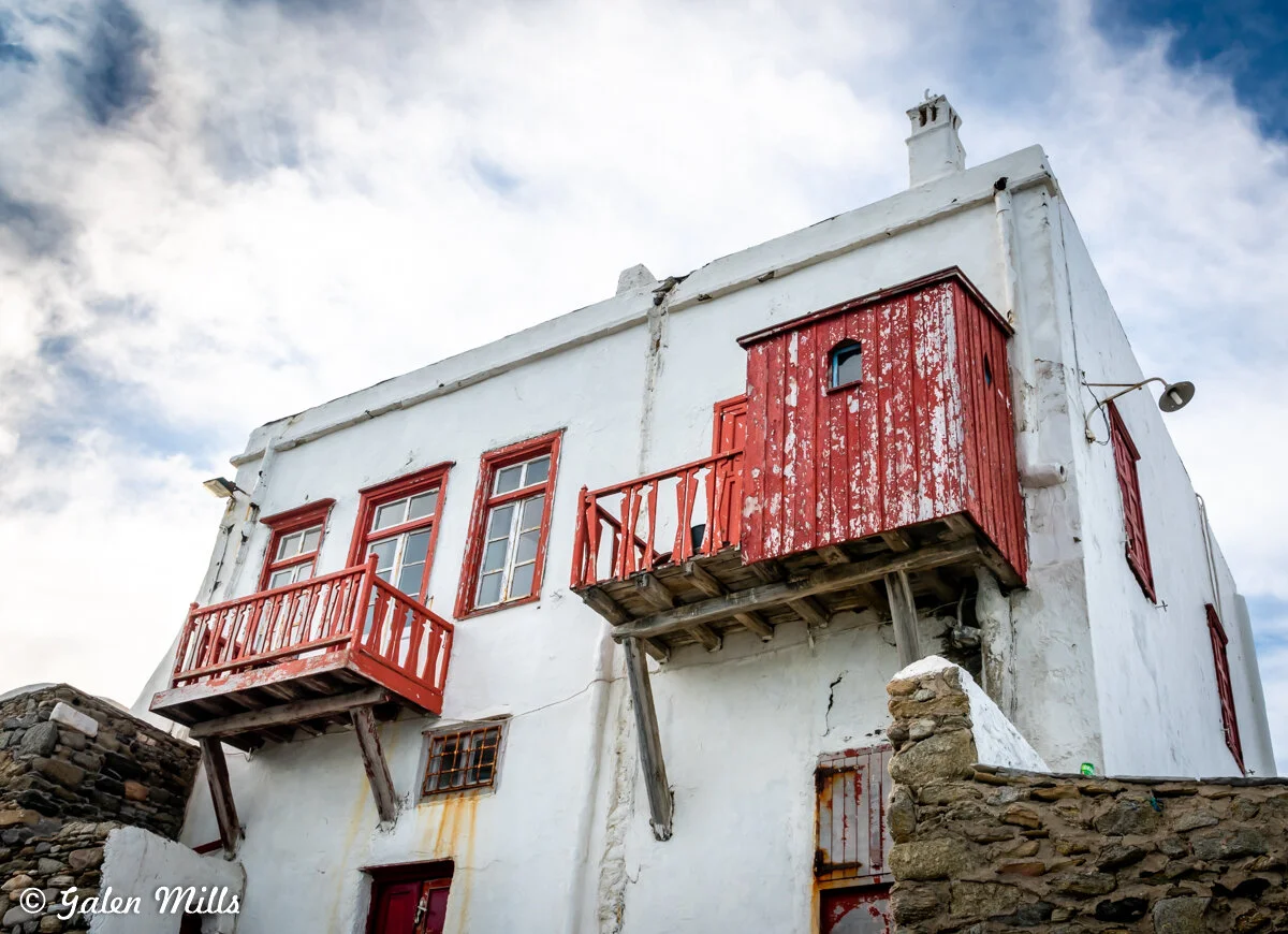 White building with red wooden balconies and shutters, weathered paint, and stone wall in front against a cloudy sky.