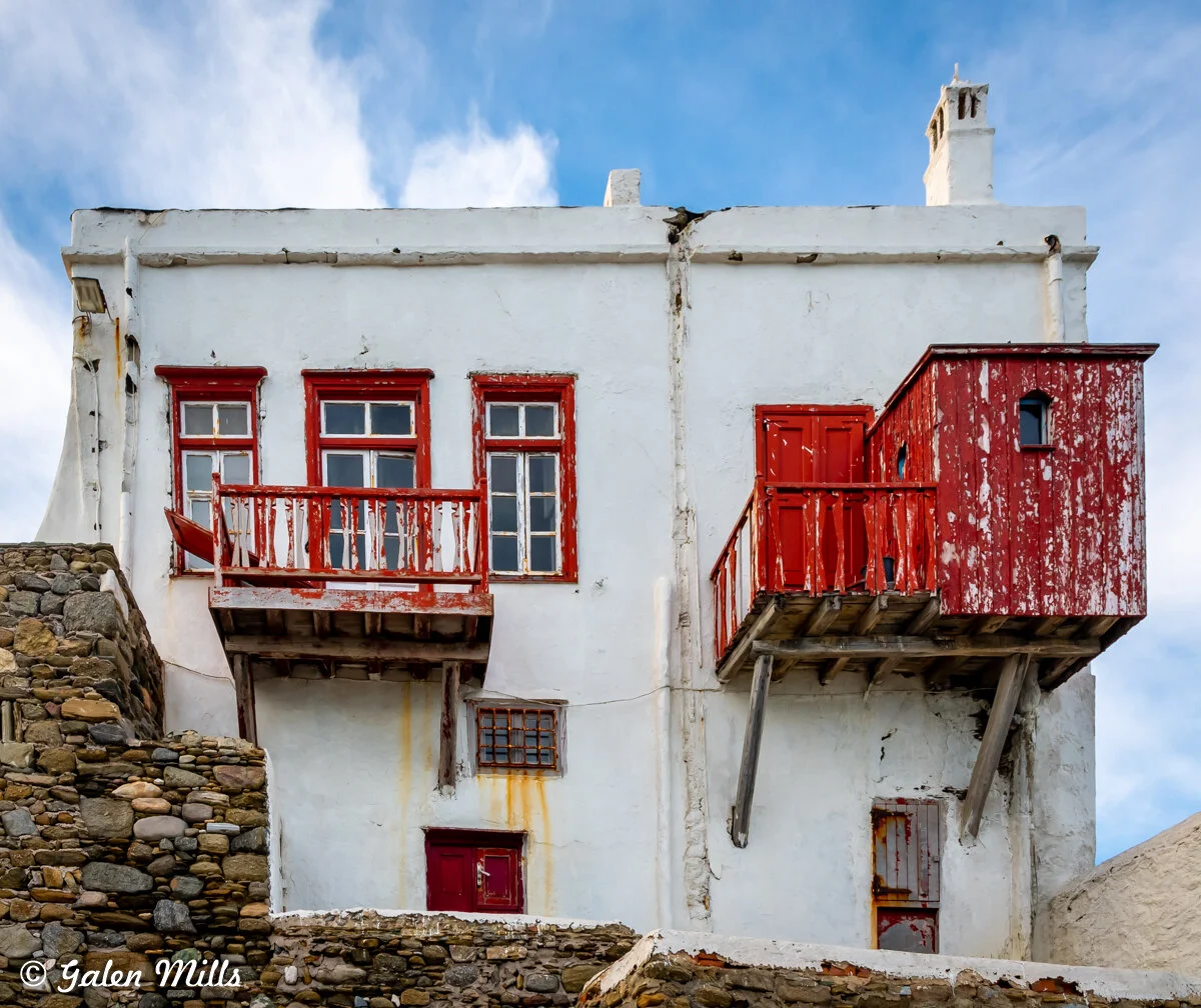 White building with red doors and windows, featuring rustic red balconies, set against a blue sky. The structure has an aged appearance with weathered wood.