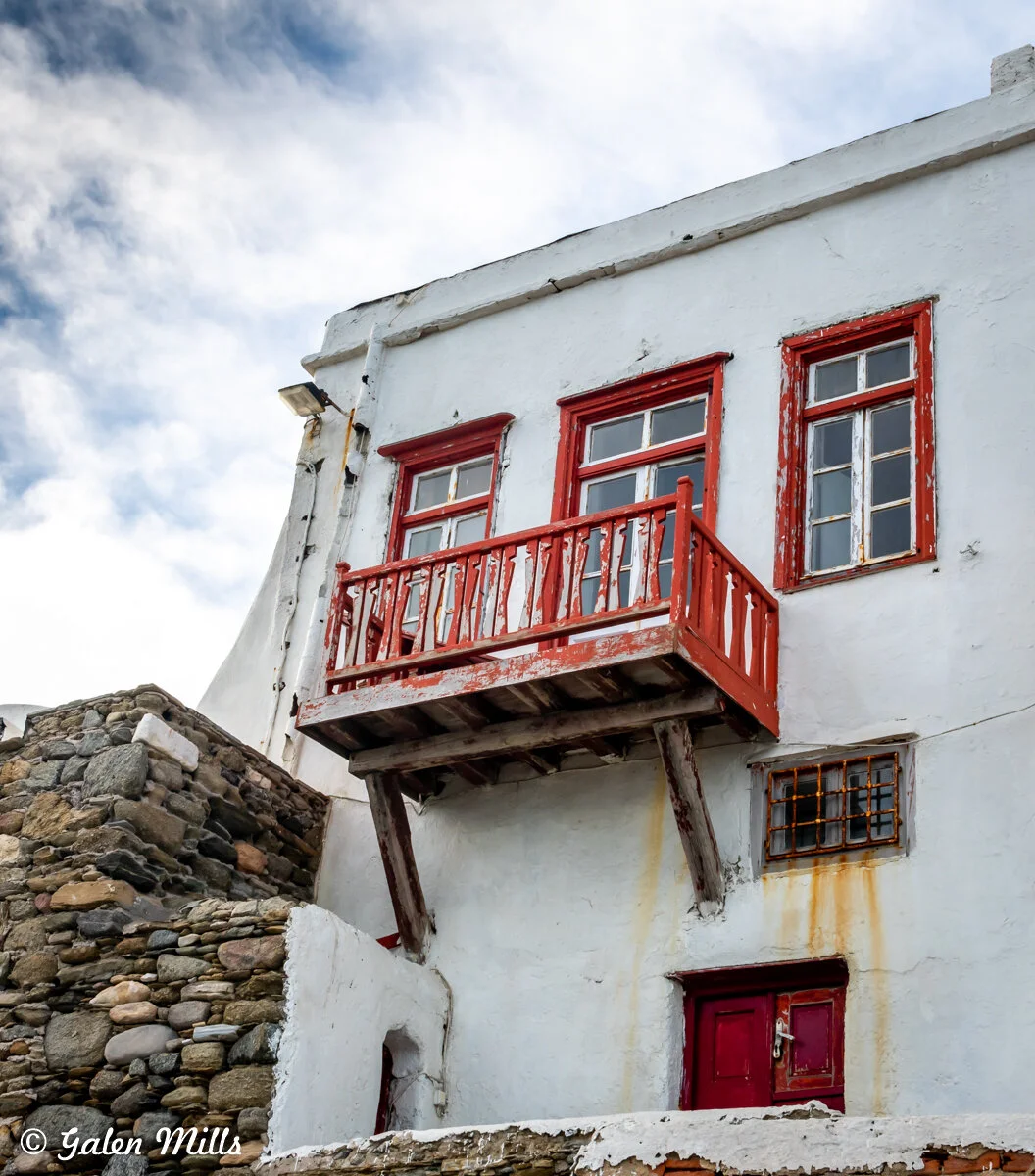 White building with red doors and windows, rustic balcony, stone wall, cloudy sky.
