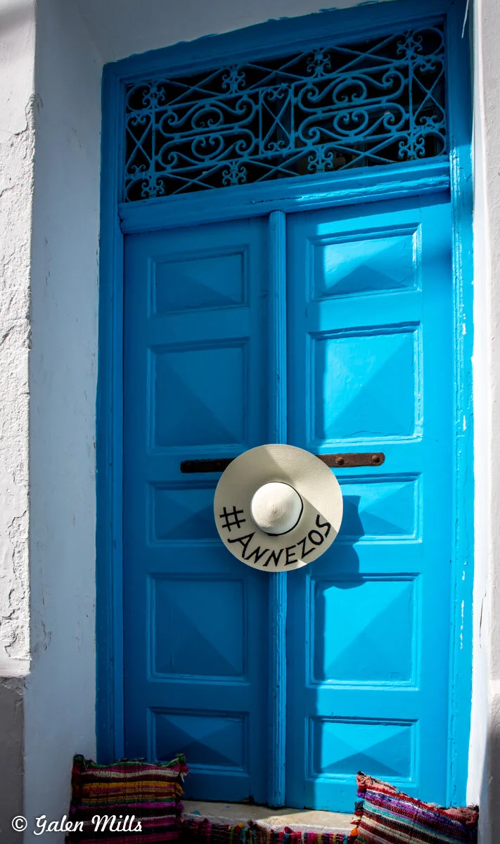 Bright blue door with decorative ironwork at the top and a straw hat with the hashtag 'Annezos' hanging on it. Colorful woven pillows are placed at the base of the door.
