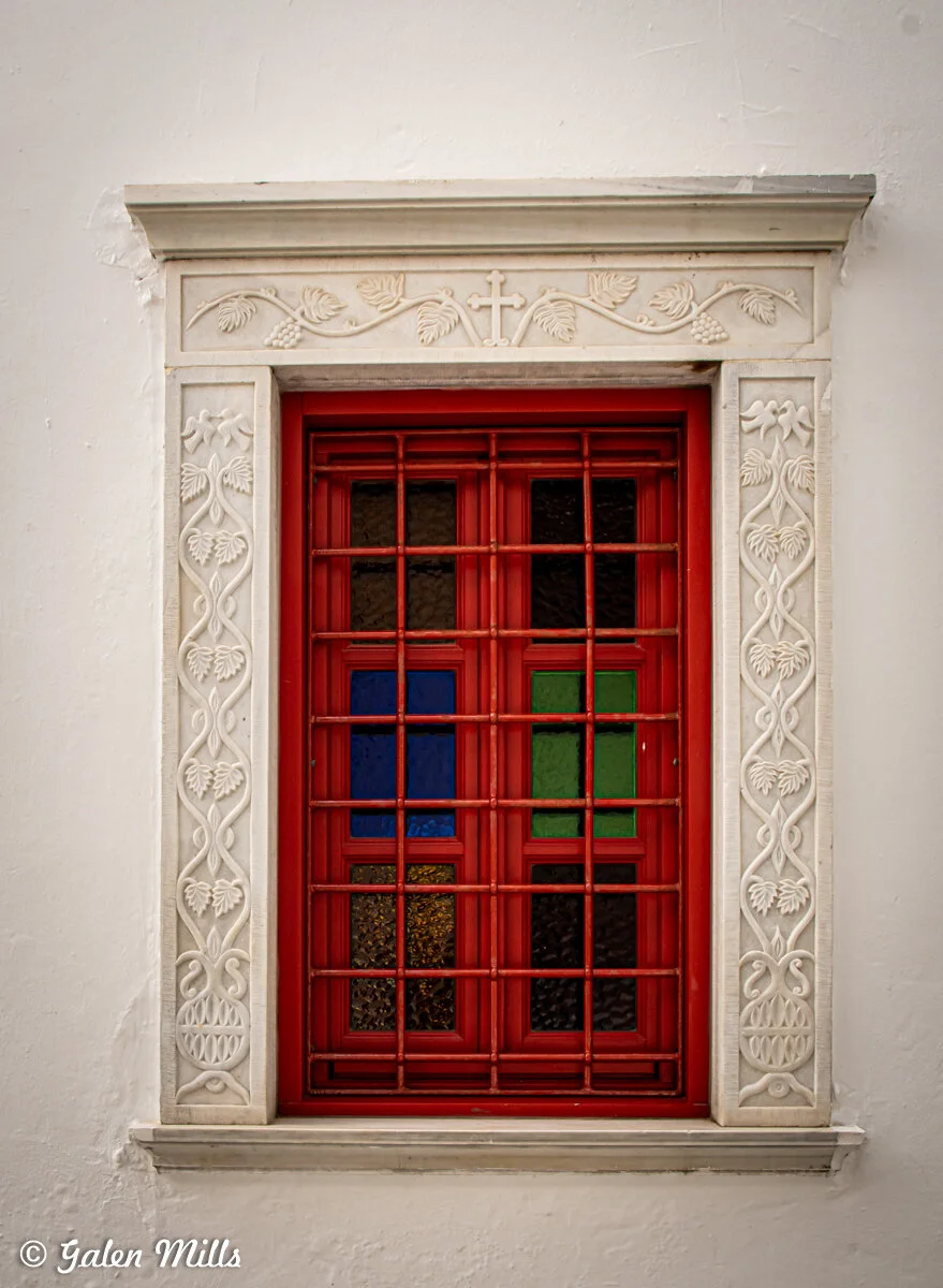 A decorative window with a red frame and metal bars, featuring colorful stained glass panels in blue, green, and yellow. The window is set in a white wall with intricate floral carvings and a cross at the top.