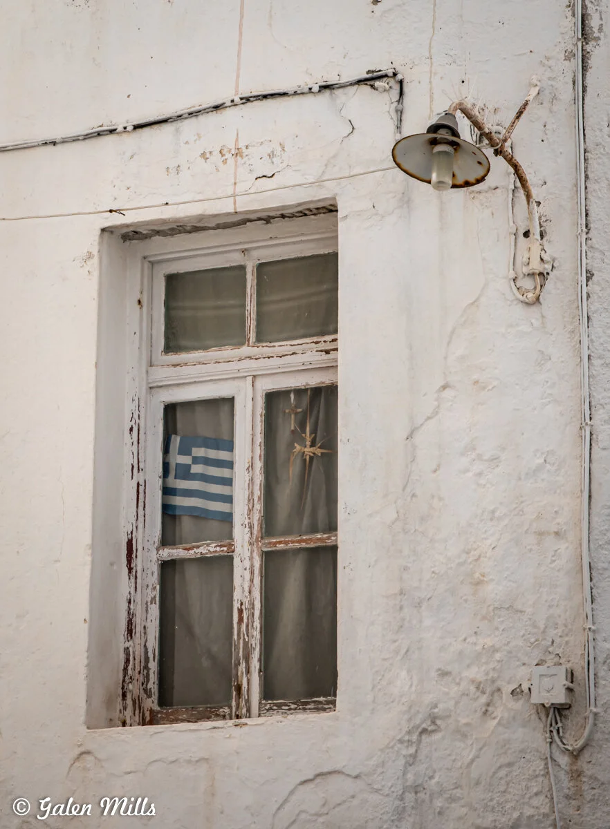 A weathered window with a Greek flag and a star ornament inside, mounted on a white textured wall. An exterior lamp is attached next to the window.