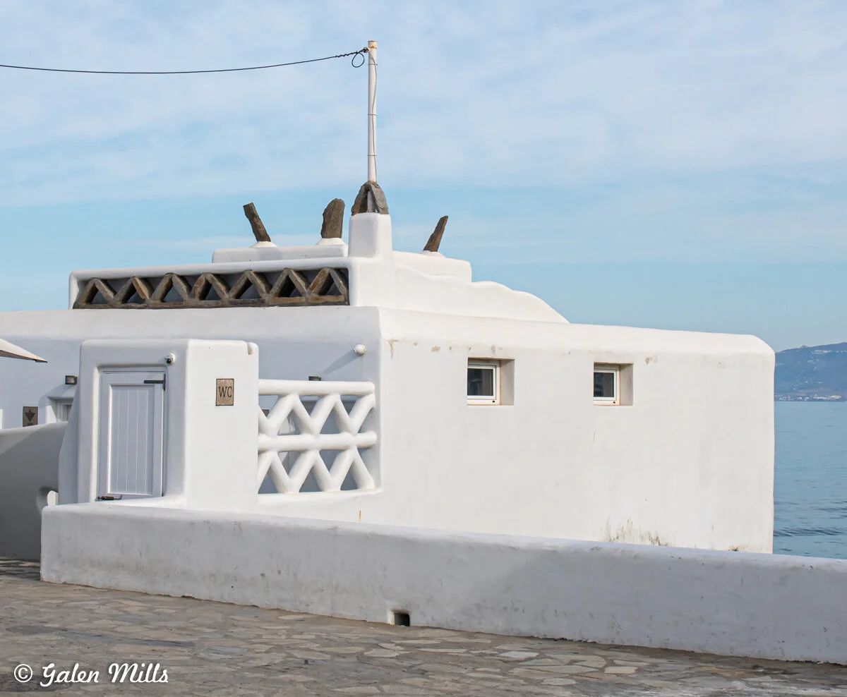 White stucco building with a WC sign, blue sky and ocean view, Mykonos style architecture.