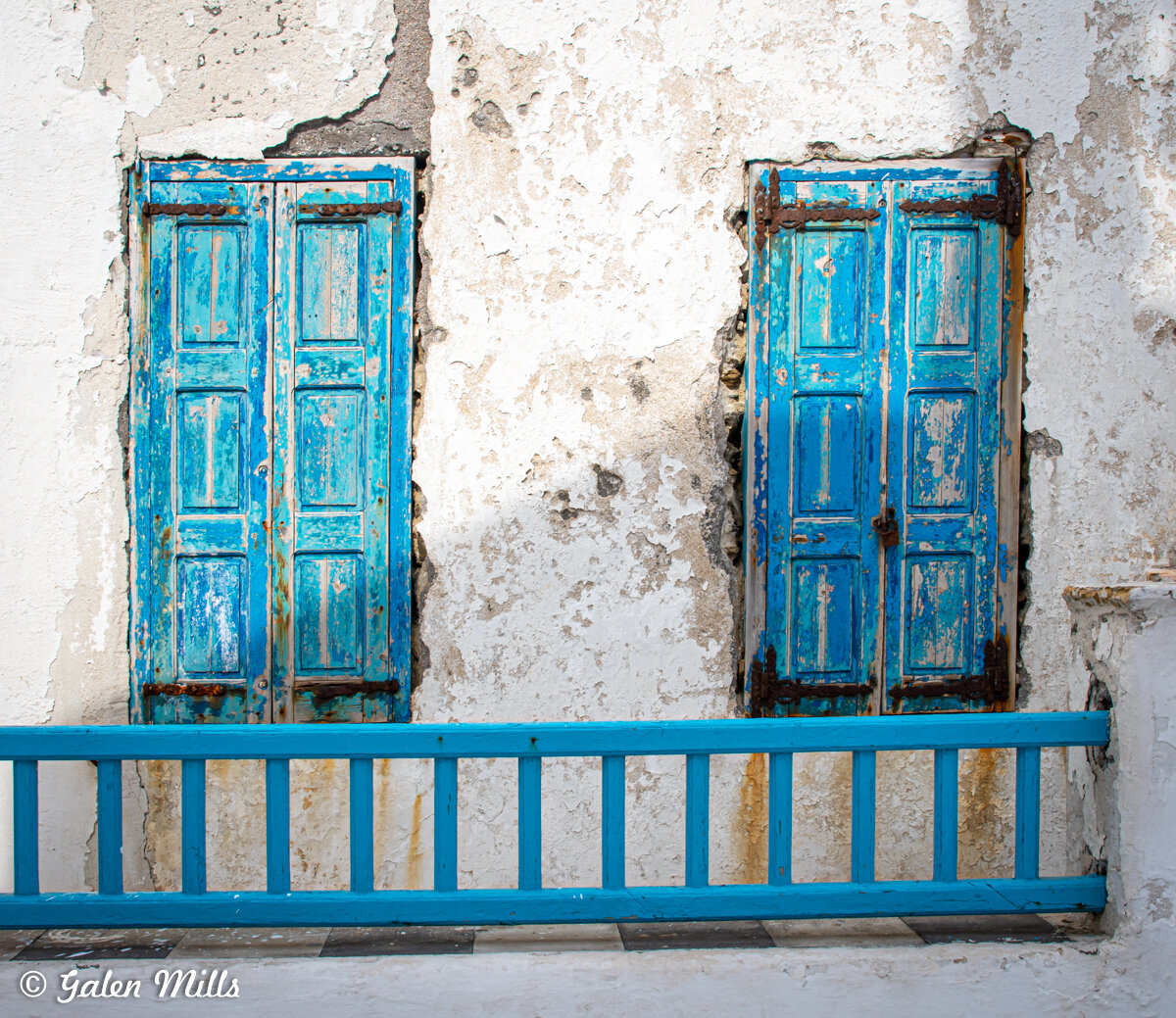 Two old blue wooden shutters on a weathered white wall with peeling paint, separated by a small blue railing.