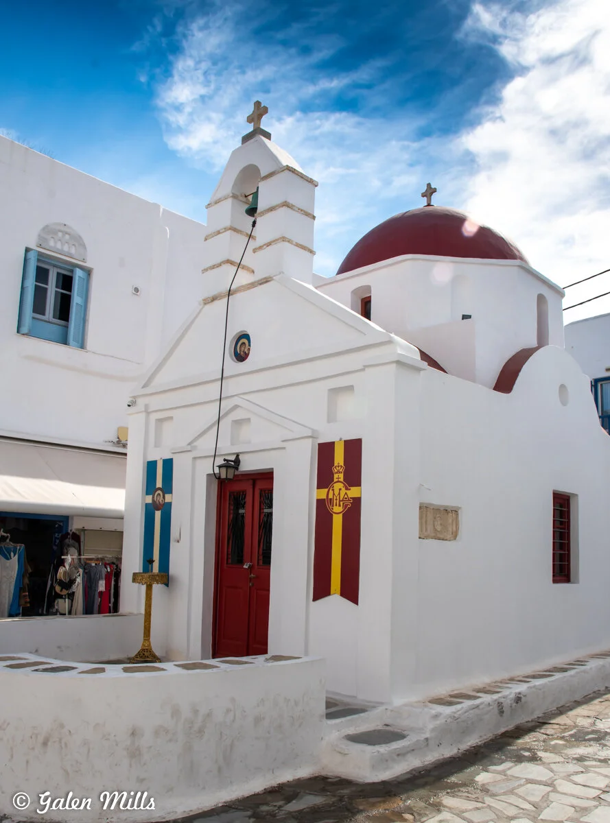 White Orthodox chapel with a red dome, cross, and flags in Mykonos, Greece under a blue sky.