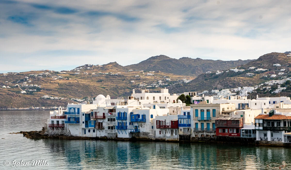 Scenic view of Little Venice in Mykonos, Greece with white buildings and colorful balconies along the waterfront, hills in the background, and calm water reflecting the structures.