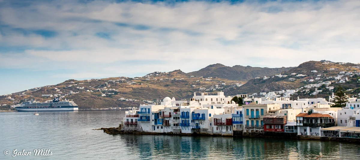 View of Mykonos with white and blue buildings along the shoreline, distant hills, and a cruise ship in the background.