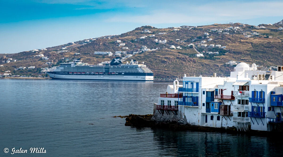 A cruise ship near the coast of Mykonos, Greece, with traditional white and blue buildings in the foreground and rugged hills in the background.
