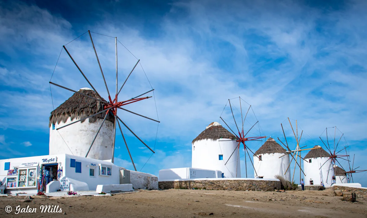 Traditional white windmills with thatched roofs in Mykonos, Greece, against a blue sky.