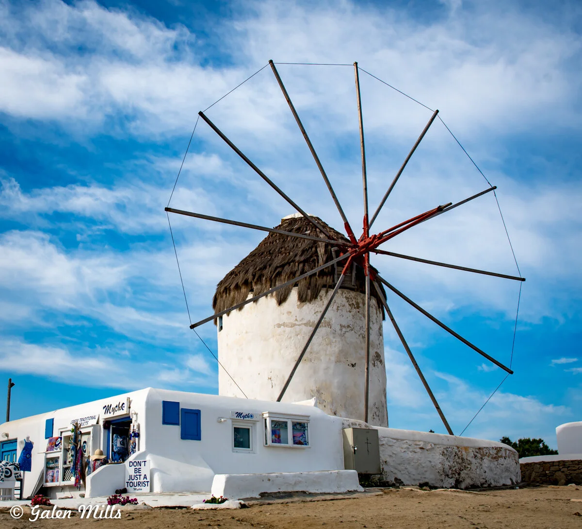 Traditional white windmill with wooden sail structure against a blue sky, next to a white building with a blue door and windows.