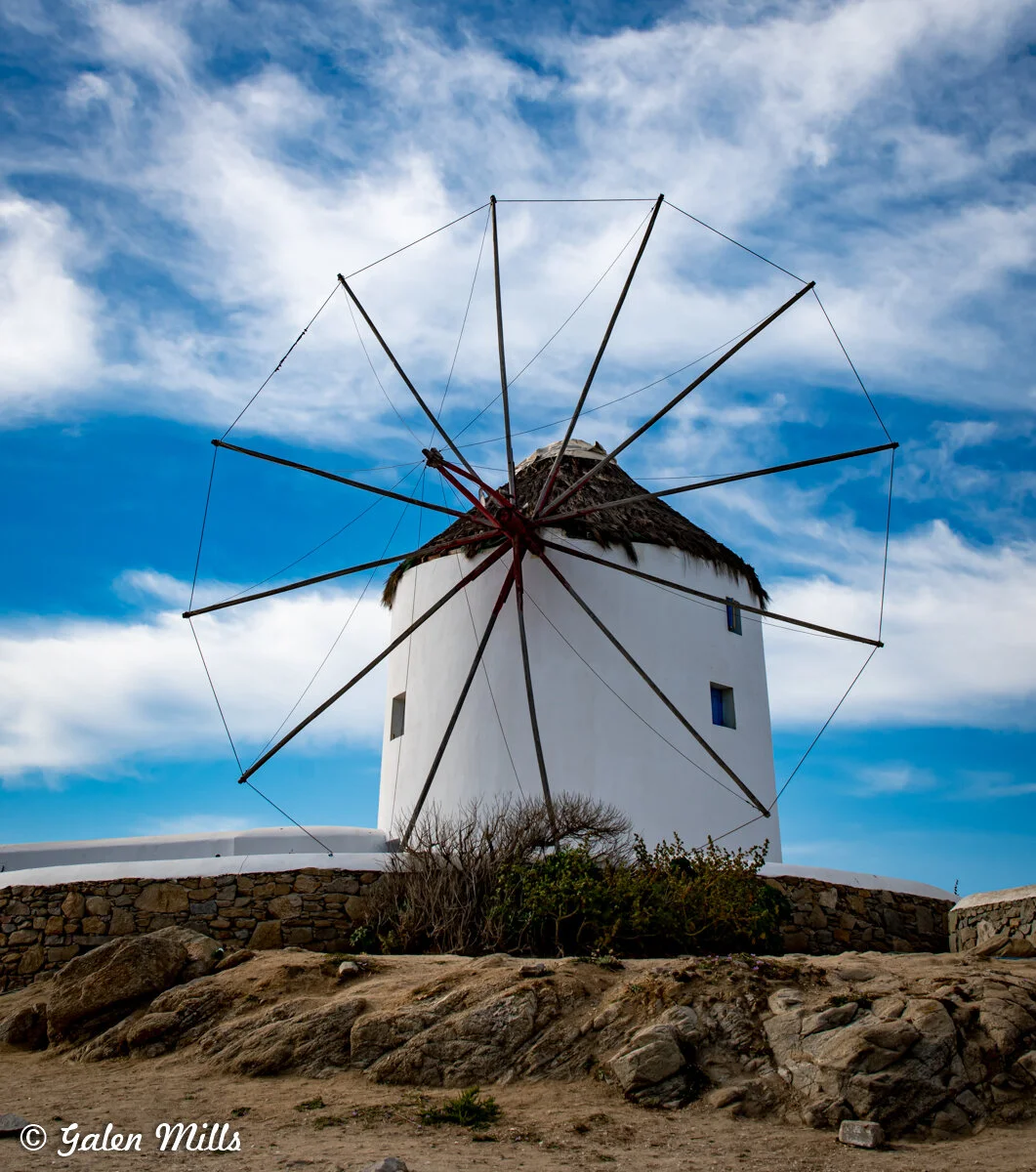 Traditional white windmill with wooden blades on a rocky landscape under a blue sky with scattered clouds.