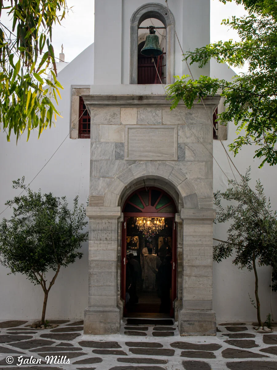 Stone church entrance with bell tower, arched doorway, and surrounding greenery.