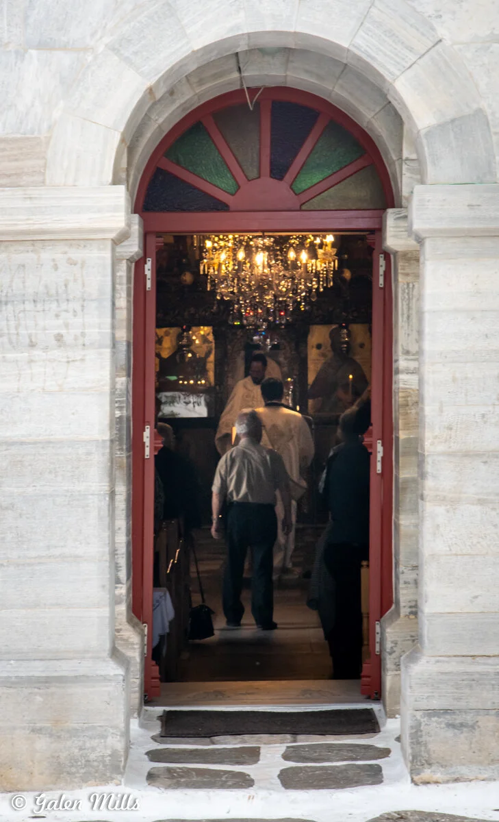 View through an open arched doorway of a stone building revealing people inside a church, with a chandelier and religious icons visible.