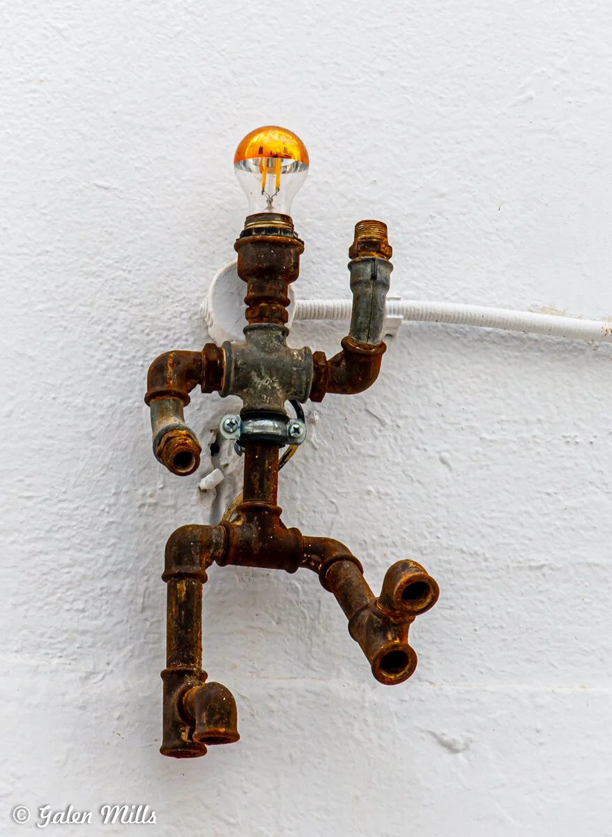 Metal sculpture of a running figure made from pipes with a lightbulb head, mounted on a white wall.