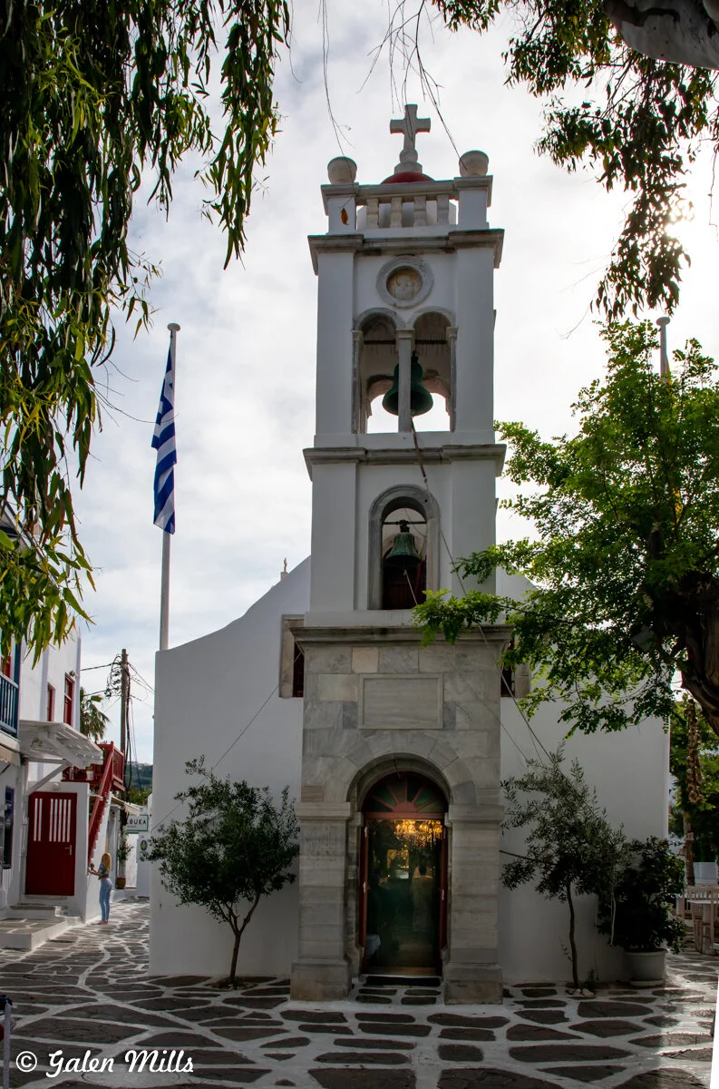 Bell tower of a Greek Orthodox church with a cross on top, surrounded by trees and a Greek flag, with a cobblestone path leading to the entrance.