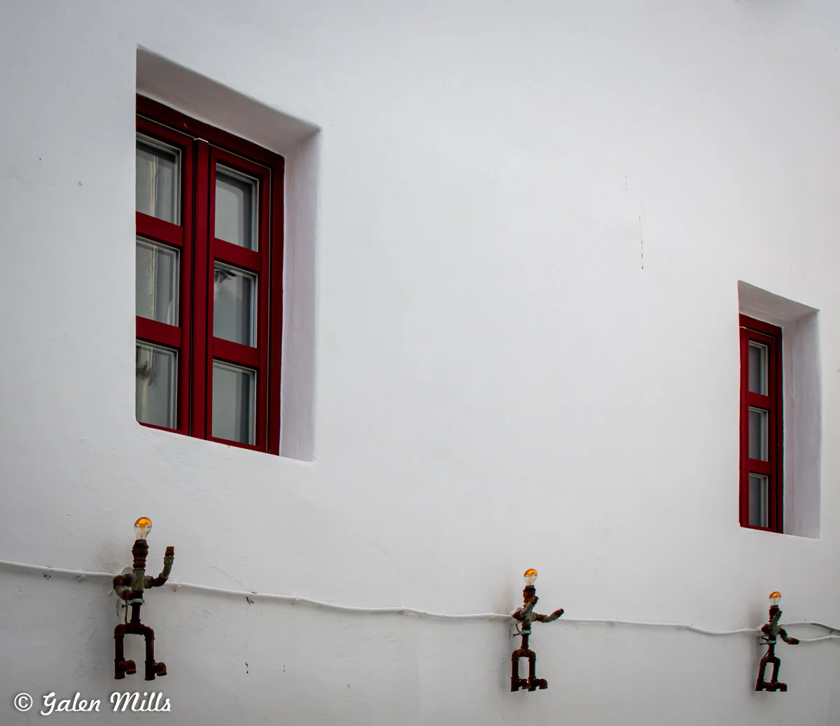White plaster wall with two red-framed windows and three pipe-shaped ornamental light fixtures.