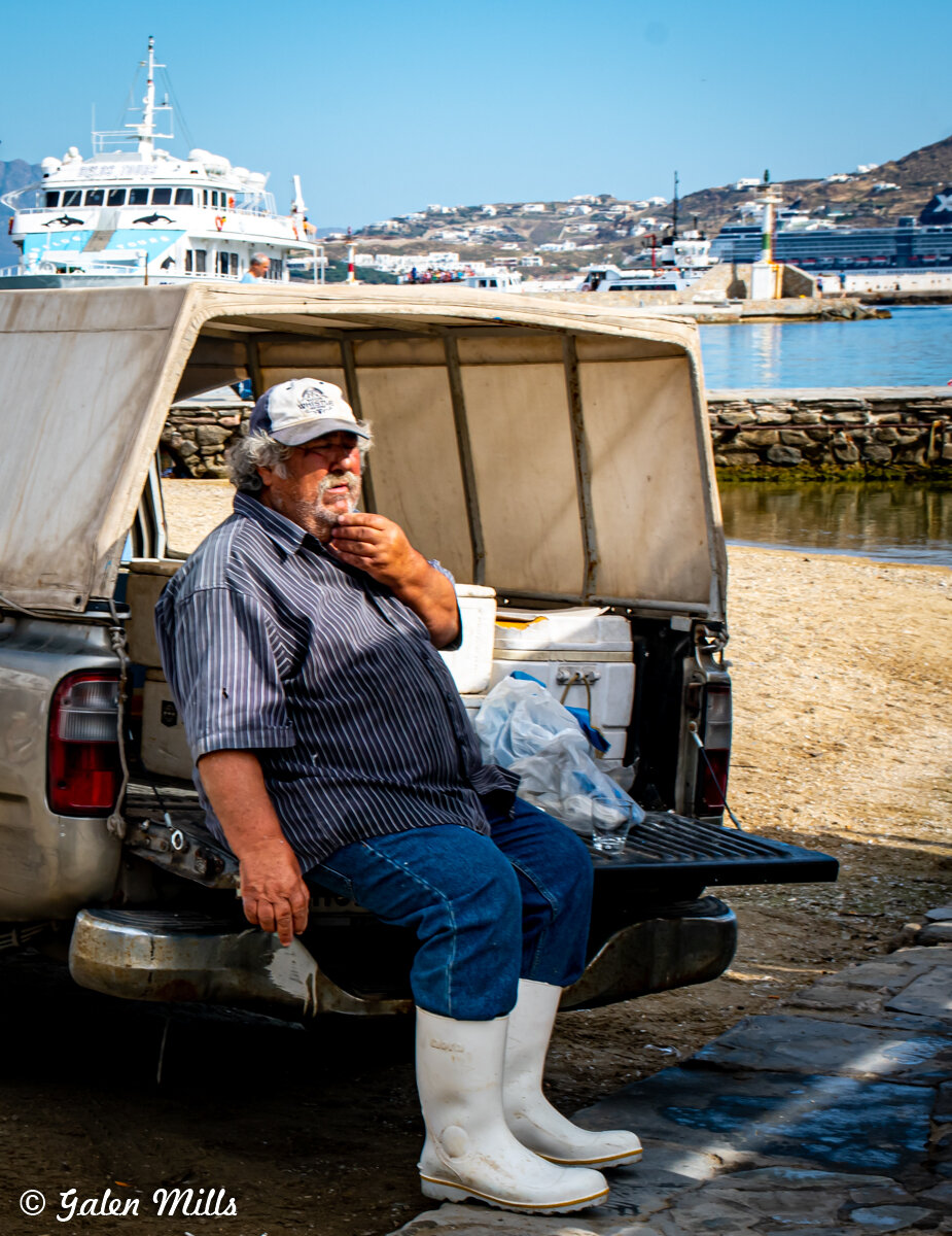 A man wearing a cap, blue jeans, white boots, and a striped shirt sits on the tailgate of a truck by the waterfront. A large boat is docked in the background, with hills and buildings in the distance.