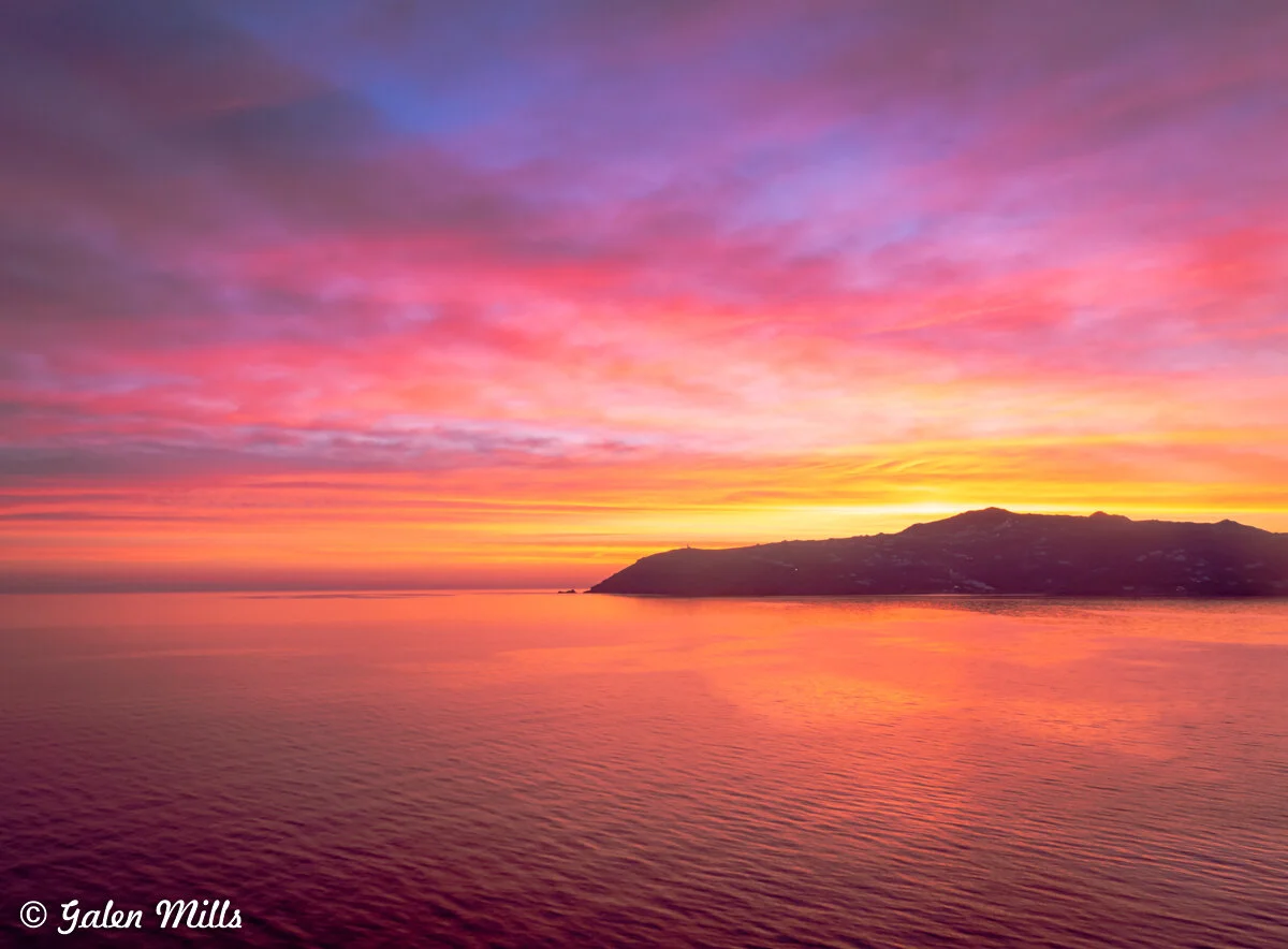 A vibrant sunset over the ocean with a silhouette of a distant coastline. The sky is filled with shades of pink, orange, and purple.
