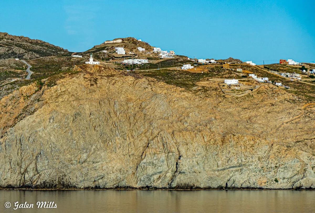 Coastal landscape with rocky cliffs and white buildings on a hillside, clear blue sky.