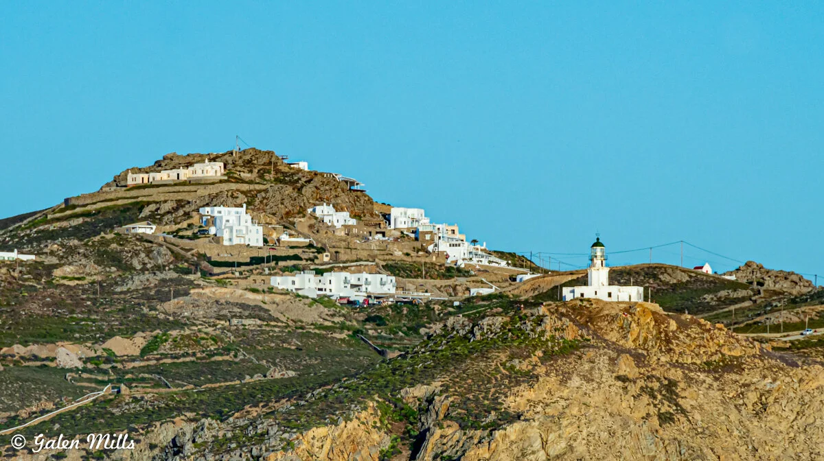 Landscape view of a hilltop village with white buildings and a lighthouse, set against a clear blue sky.
