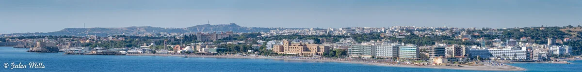 Panoramic view of a coastal city with buildings along the shoreline, clear blue sea, and a distant hilly landscape.