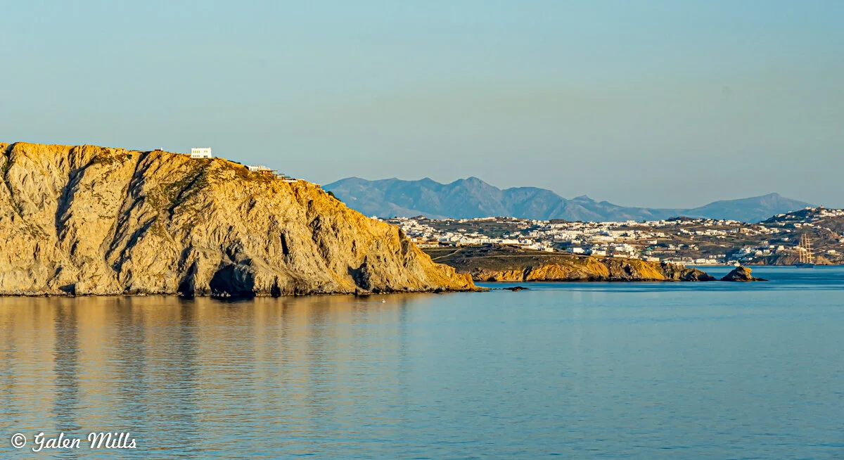 Coastal landscape with rocky cliffs and distant town, calm sea reflecting golden light, and mountain range under a clear sky.