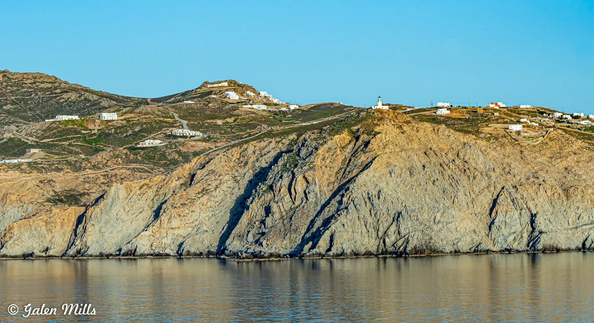 Landscape of a rocky coastline with scattered white buildings and a lighthouse under a clear blue sky.