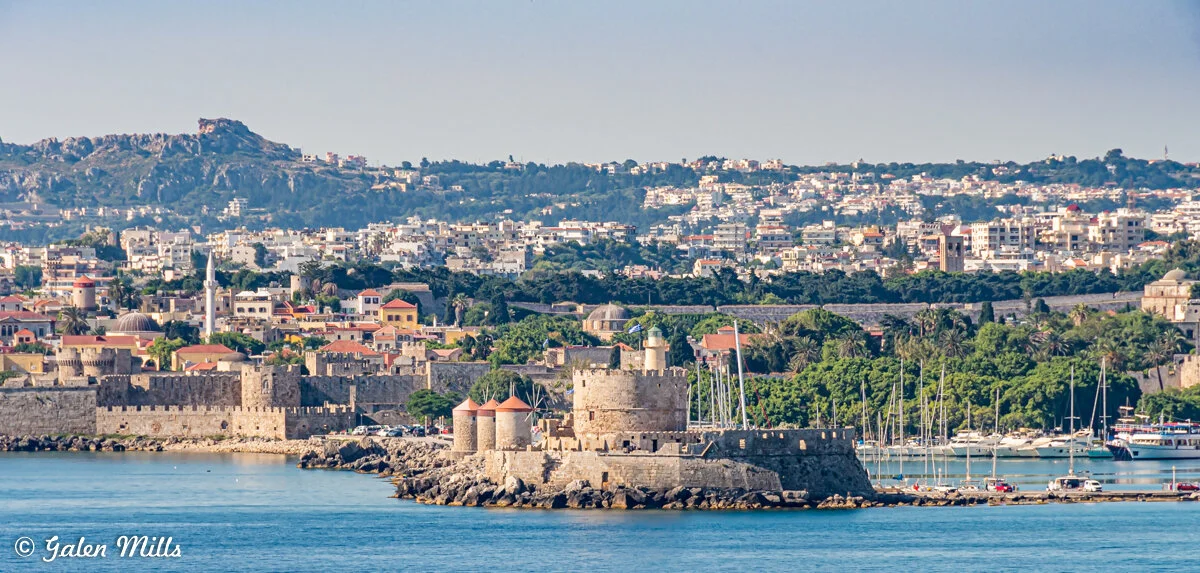 View of medieval castle and fortifications on the coastline of Rhodes, Greece, with mountain backdrop and cityscape.