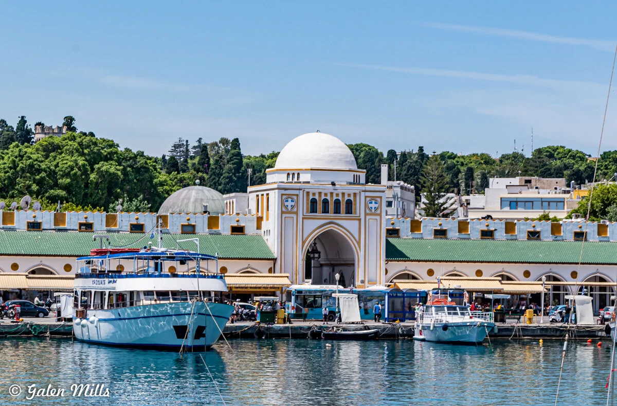 View of the port area with moored boats and a distinctive domed building, with a backdrop of trees and urban architecture under a clear blue sky.
