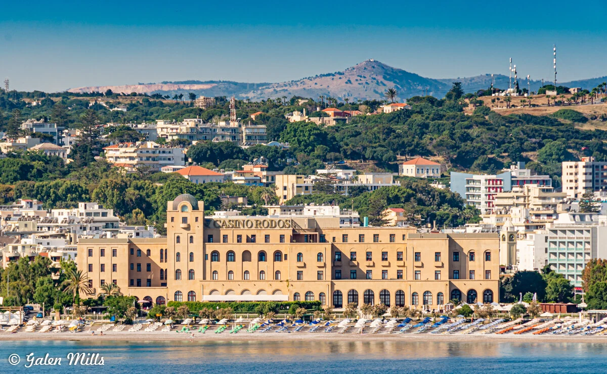 Scenic view of Casino Rodos building with beachfront and surrounding town, nestled against a backdrop of lush green hills and distant mountains in Rhodes, Greece.