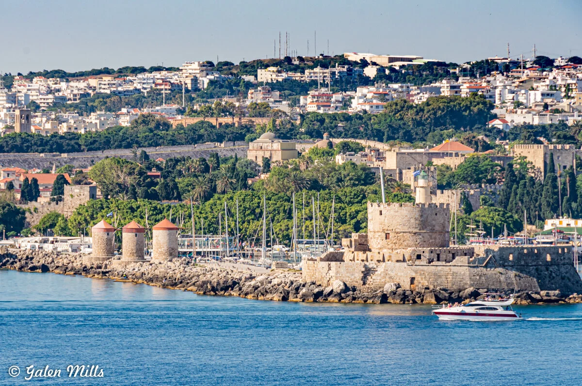 Scenic view of the historic port of Rhodes, Greece, featuring medieval stone fortress walls, windmills, and buildings against a city and lush greenery background, with a boat on the sea.