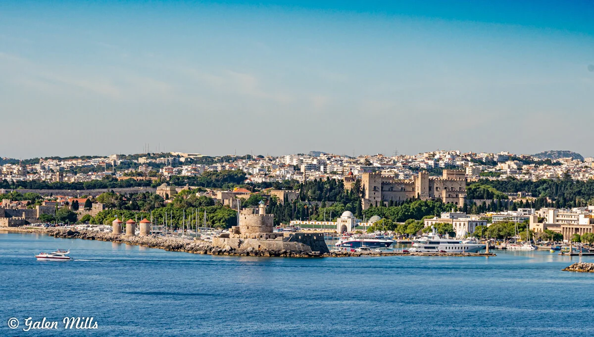 View of Rhodes, Greece from the sea, featuring the medieval Palace of the Grand Master, a lighthouse, windmills, and a marina with yachts.