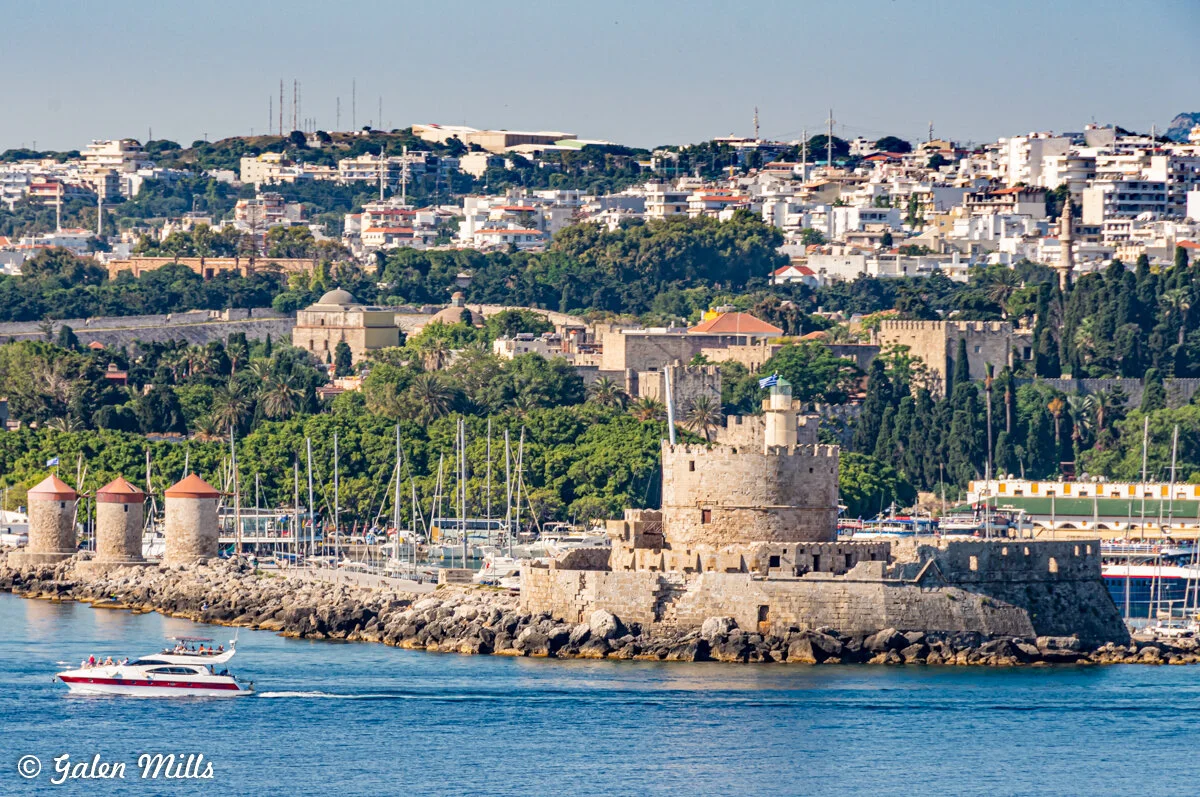 View of Rhodes harbor with historic fortifications, windmills, and a boat on the water.