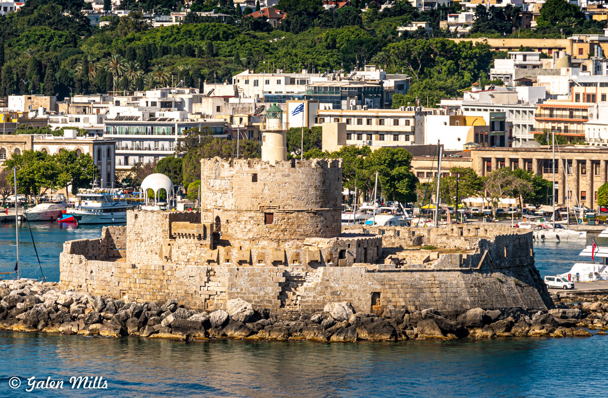 Medieval fortress on a waterfront with city buildings in the background, featuring a cylindrical tower and stone walls in a harbor setting, surrounded by boats and greenery.