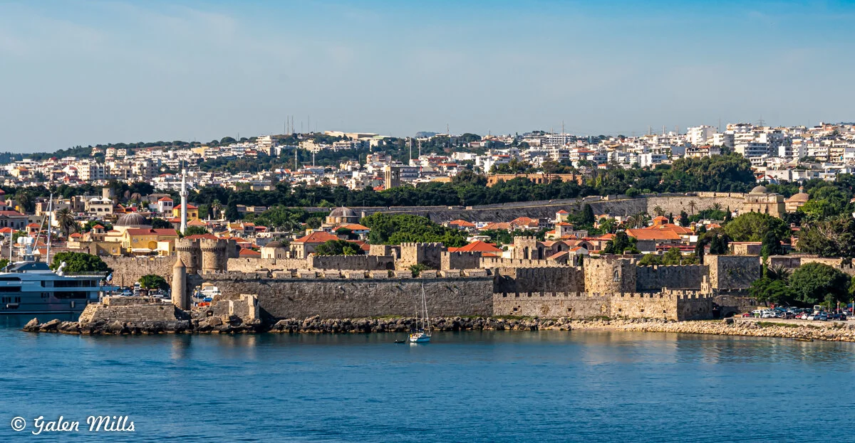 Panoramic view of the medieval city of Rhodes with stone walls, towers, and historical buildings, located by the sea, with a modern cityscape in the background under a blue sky.