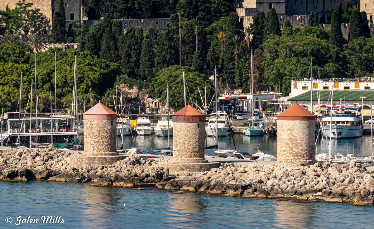 Stone windmills on the harbor with sailboats, surrounded by green trees and old buildings.